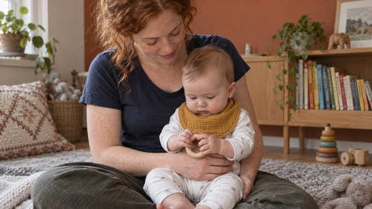 A red-haired parent sitting with an awake baby wearing a dry bib in a bright terracotta-accented room.