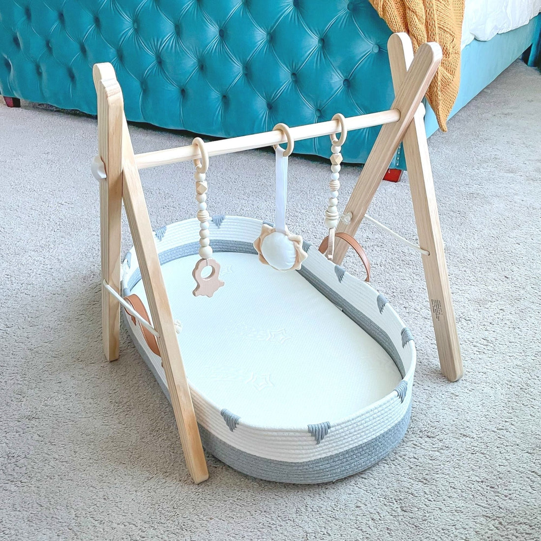 Freestanding wooden baby gym with hanging toys above a gray and white Vesta Baby changing basket on the floor.
