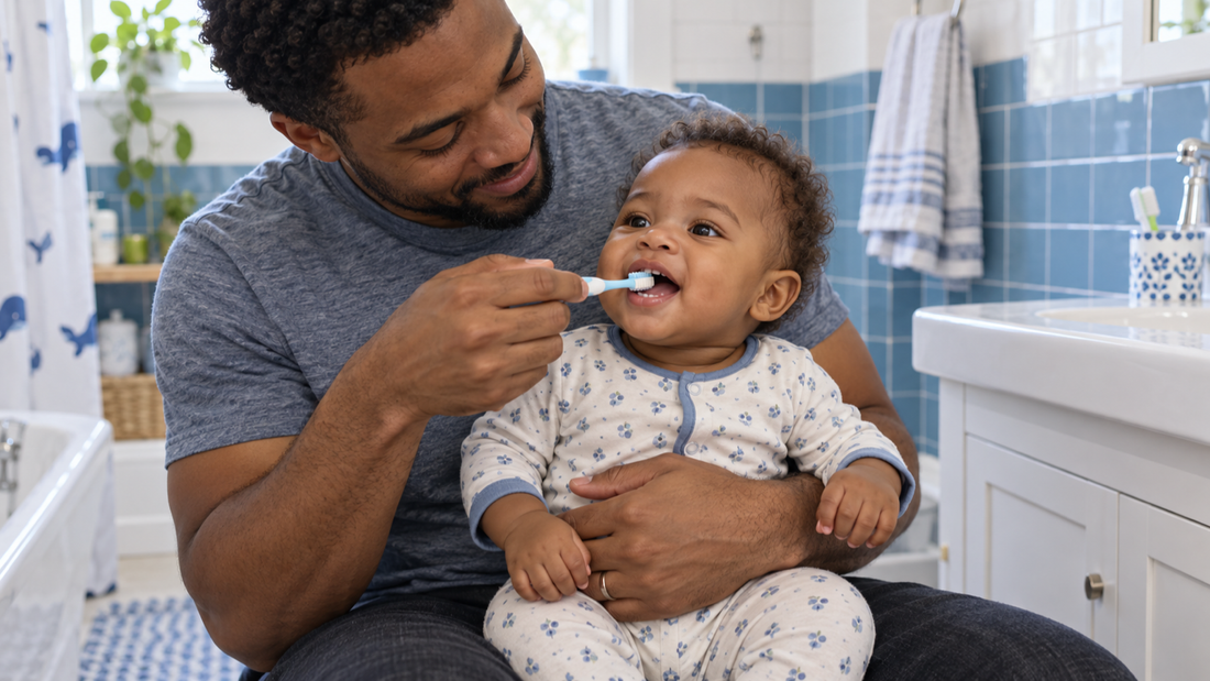 A father holding his baby securely on his lap while gently brushing the baby’s teeth in a blue-and-white bathroom.